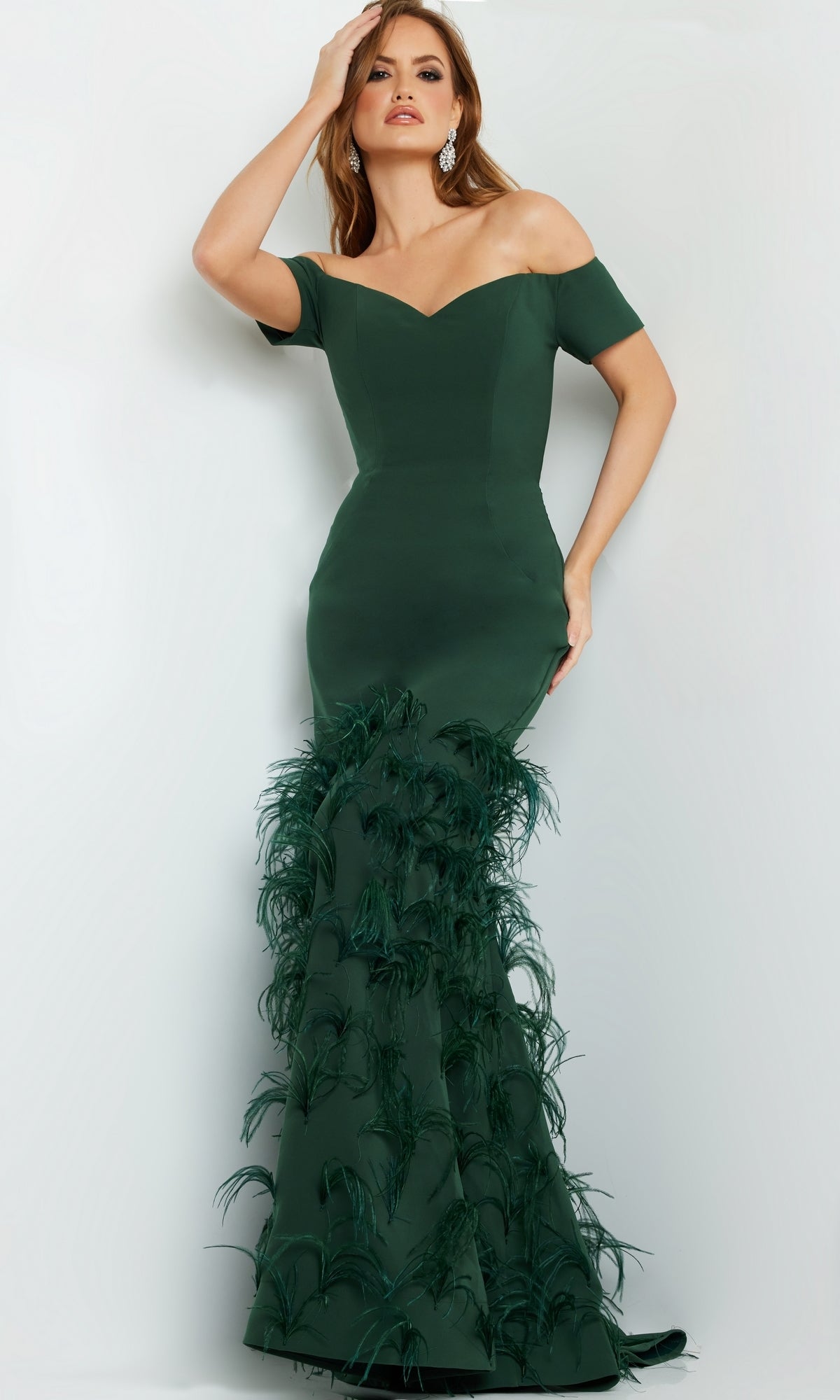 A woman stands against a plain light background wearing the Jovani 08384 Off-Shoulder Feather Mermaid Gown, a fitted dark green evening dress with feather detailing on the lower half.